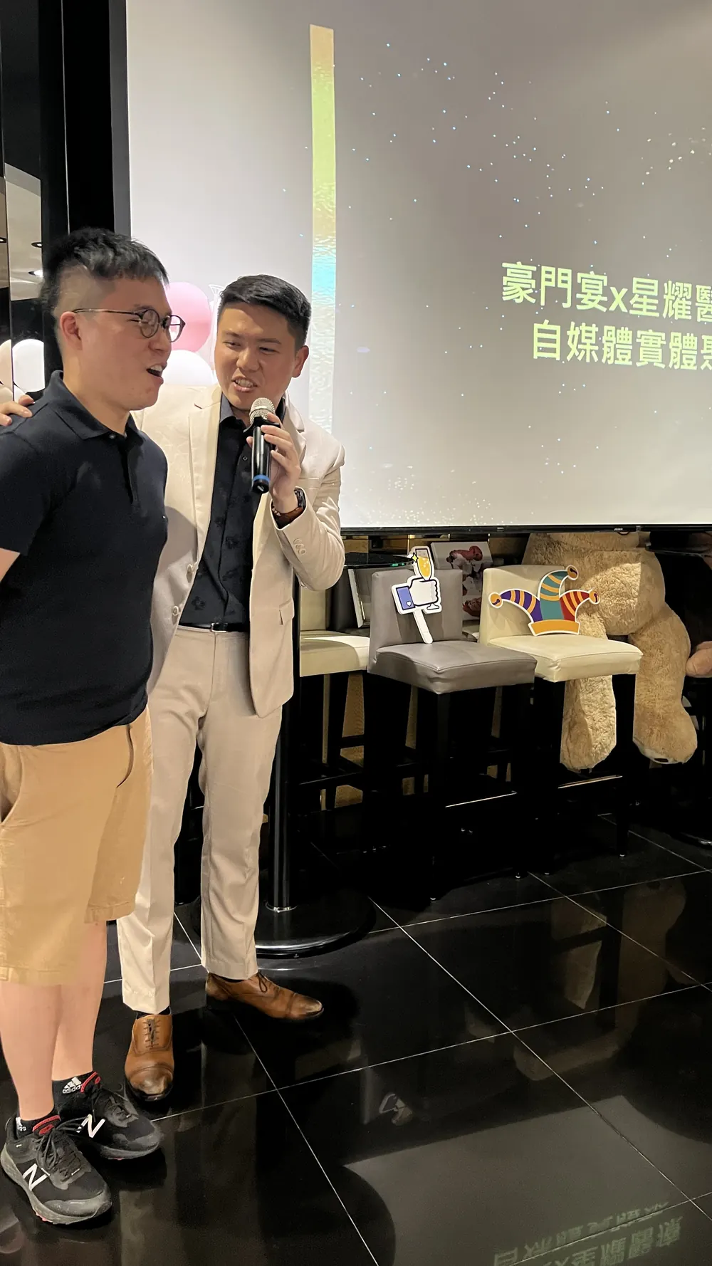 一位穿白色西裝外套的與會者手持麥克風，旁邊站著另一位與會者
