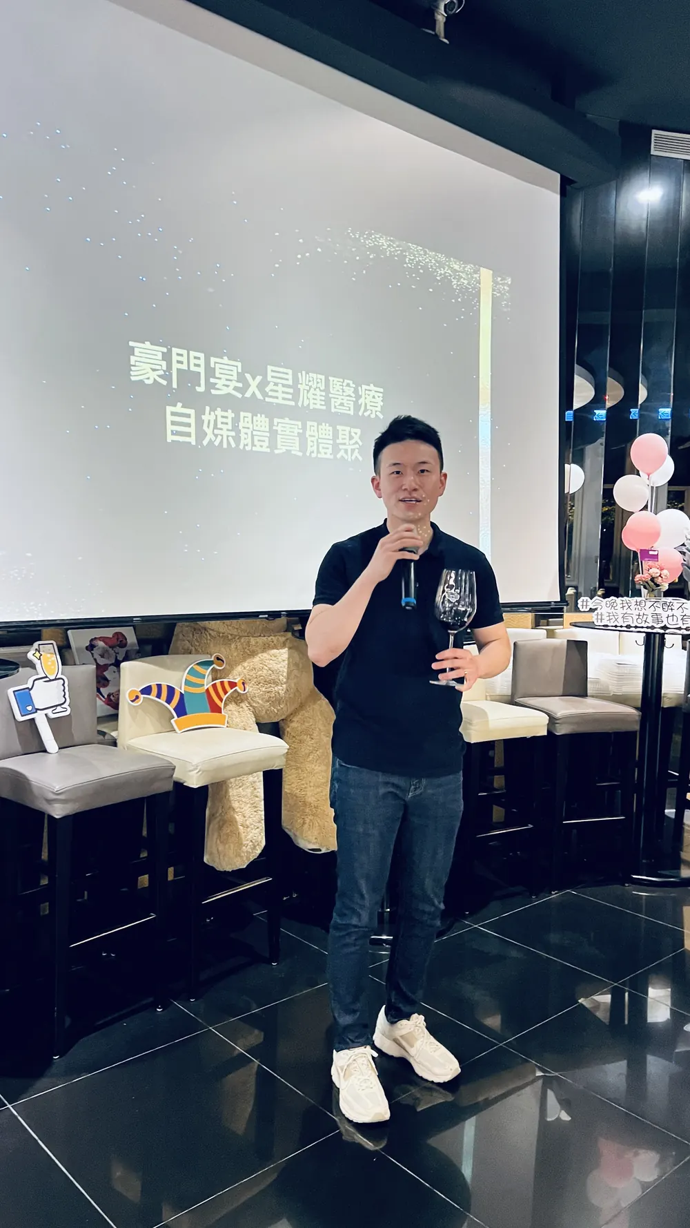 一位穿深藍色上衣的與會者手持麥克風與酒杯發言