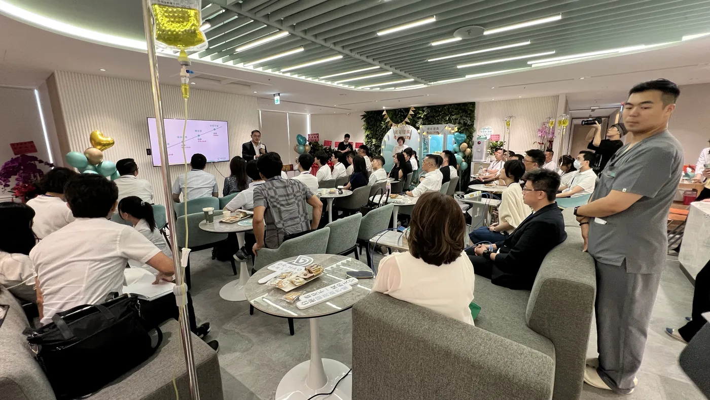 康博診所大廳坐滿與會醫師，前方電視螢幕準備開場，右側一位工作人員站著