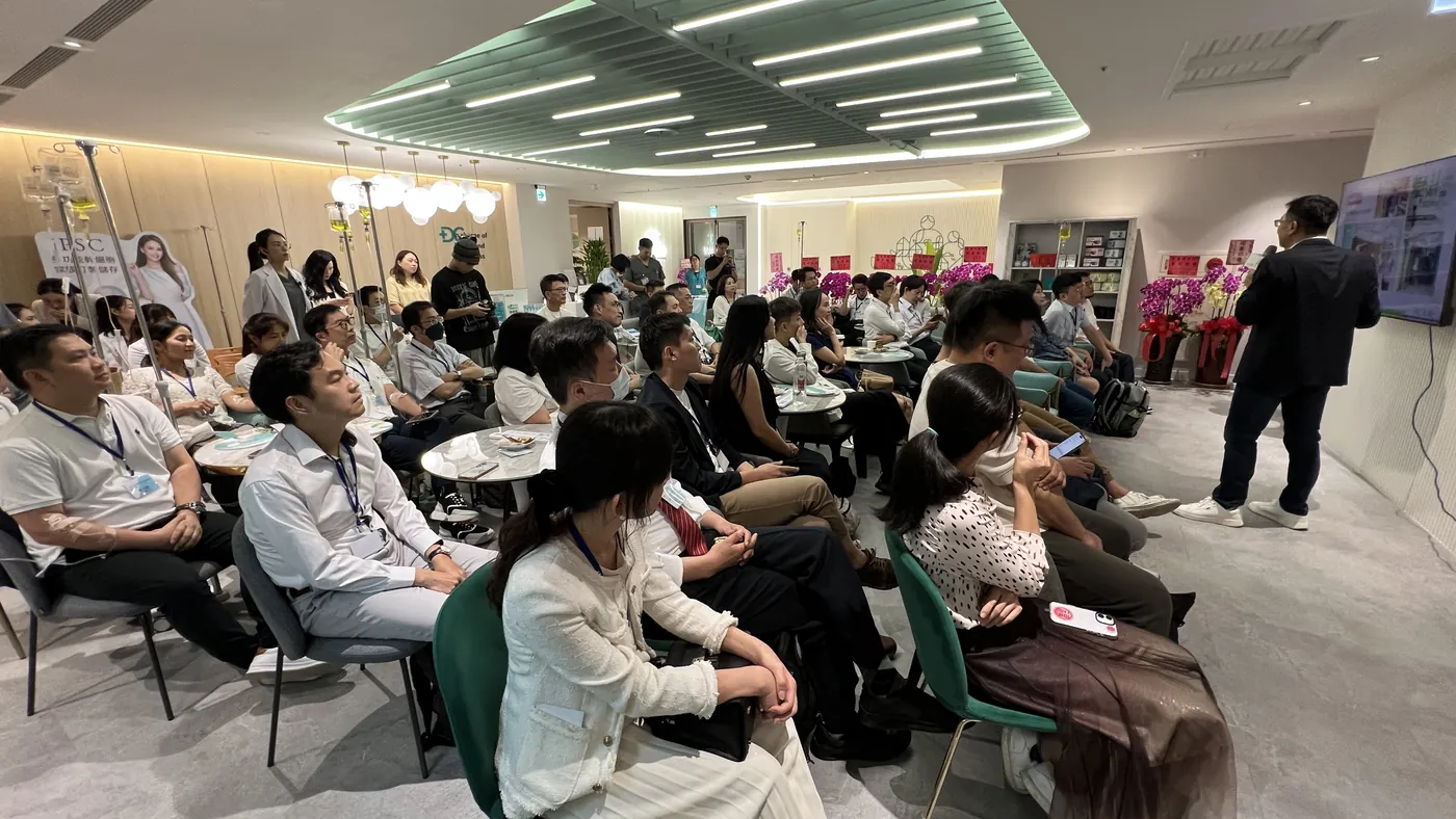 與會醫師面向前方聽講，前方有講者站在螢幕旁，右側有攝影機架設