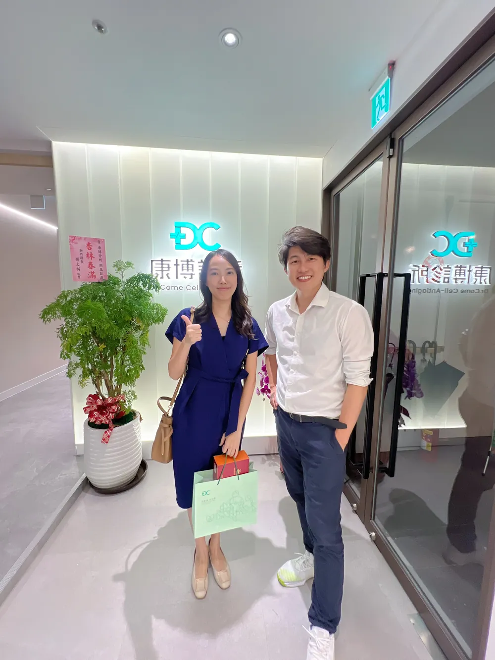 劉麗雪醫師與張益豪醫師在招牌前合影，劉醫師提著綠色禮袋比讚