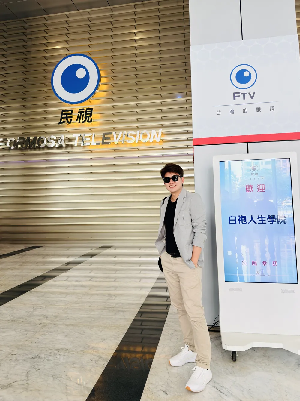 張益豪醫師站在民視大廳標誌牆前，旁邊的電子立牌顯示「歡迎 白袍人生學院 蒞臨參訪」