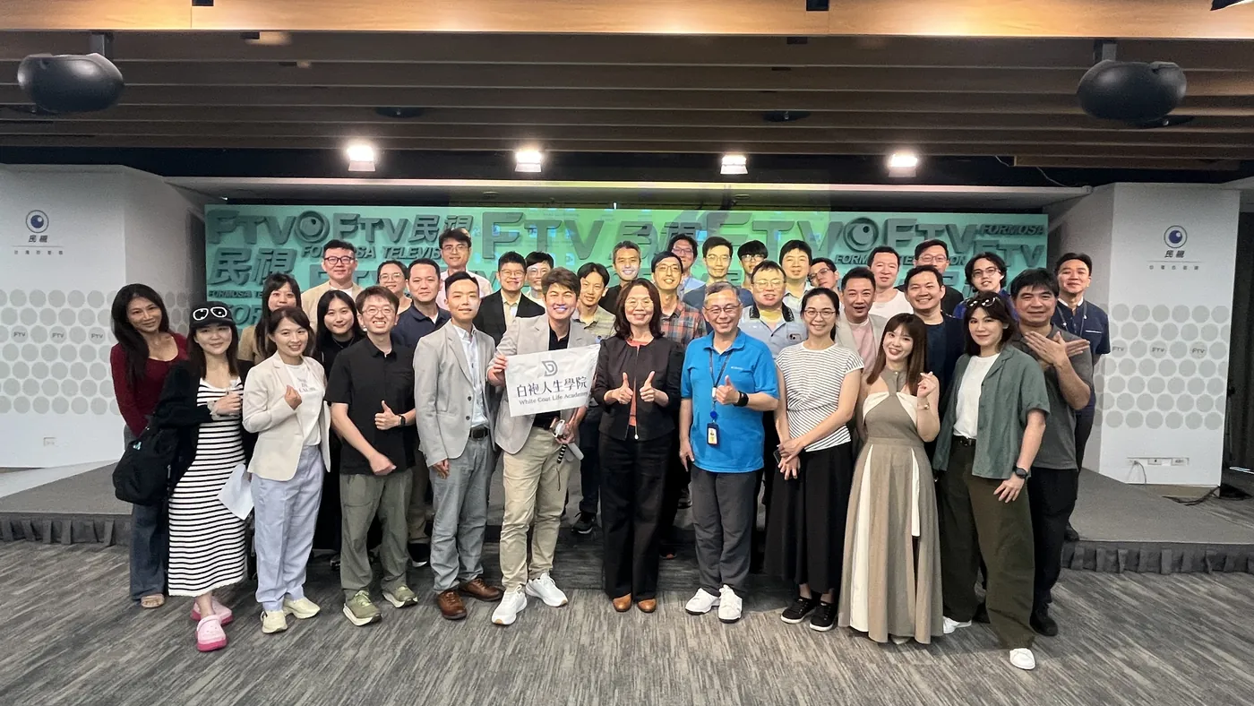 參訪醫師與民視主管在簡報室的民視背板前大合照，前排拿著白袍人生學院的旗子