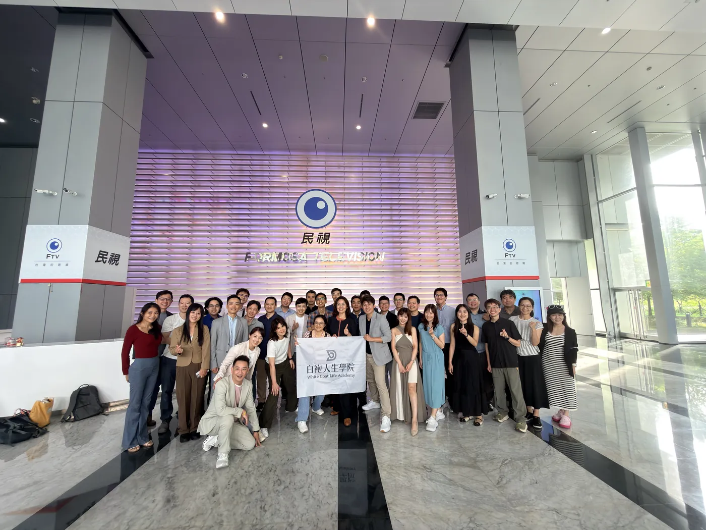 參訪醫師在民視大廳淡紫色的標誌牆前拉著白袍人生學院旗子大合照